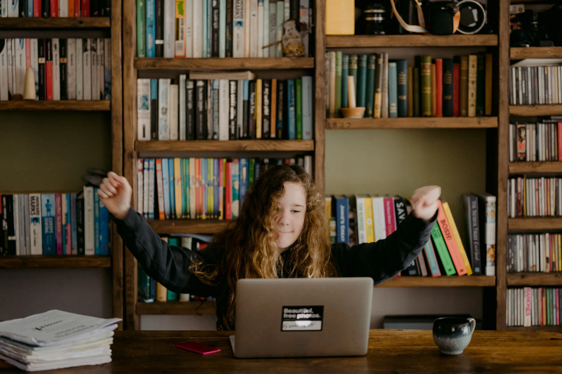 Ein Mädchen mit lockigem Haar streckt triumphierend die Arme am Schreibtisch vor einem Laptop aus, umgeben von Bücherregalen, da sie stolz auf ihren hervorragenden Lernstil ist.