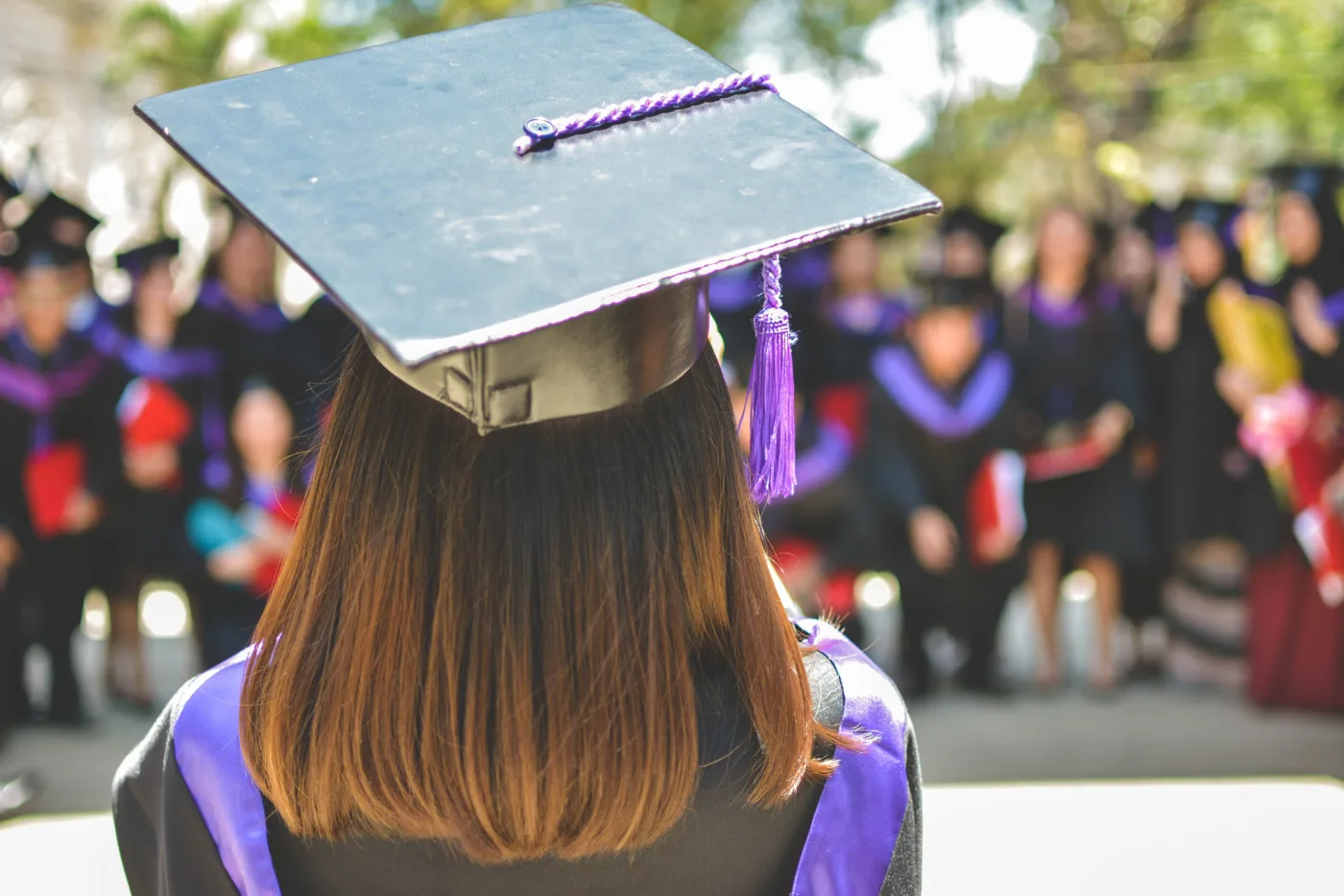 Eine Absolventin steht mit dem Rücken zur Kamera, ihr Hut deutlich sichtbar, während sie vor einer Gruppe von Absolventen im Hintergrund steht, die ebenso traditionelle akademische Kleidung tragen und ihre Lernziele erreicht haben.