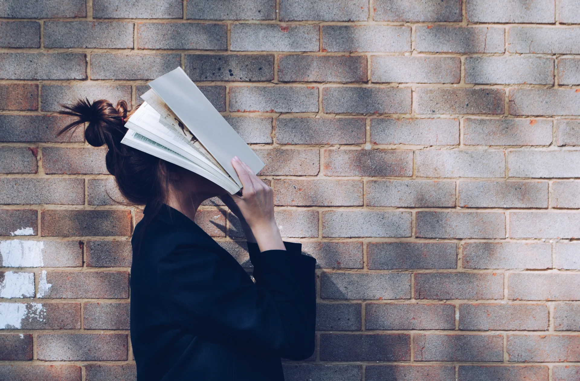 Schulstress – Eine Person hält ein aufgeschlagenes Buch vor ihr Gesicht, das sie teilweise verdeckt, vor einer Backsteinmauer, was auf das Konzept der Wissensvertiefung oder die Vermeidung von Ablenkungen hindeuten könnte.