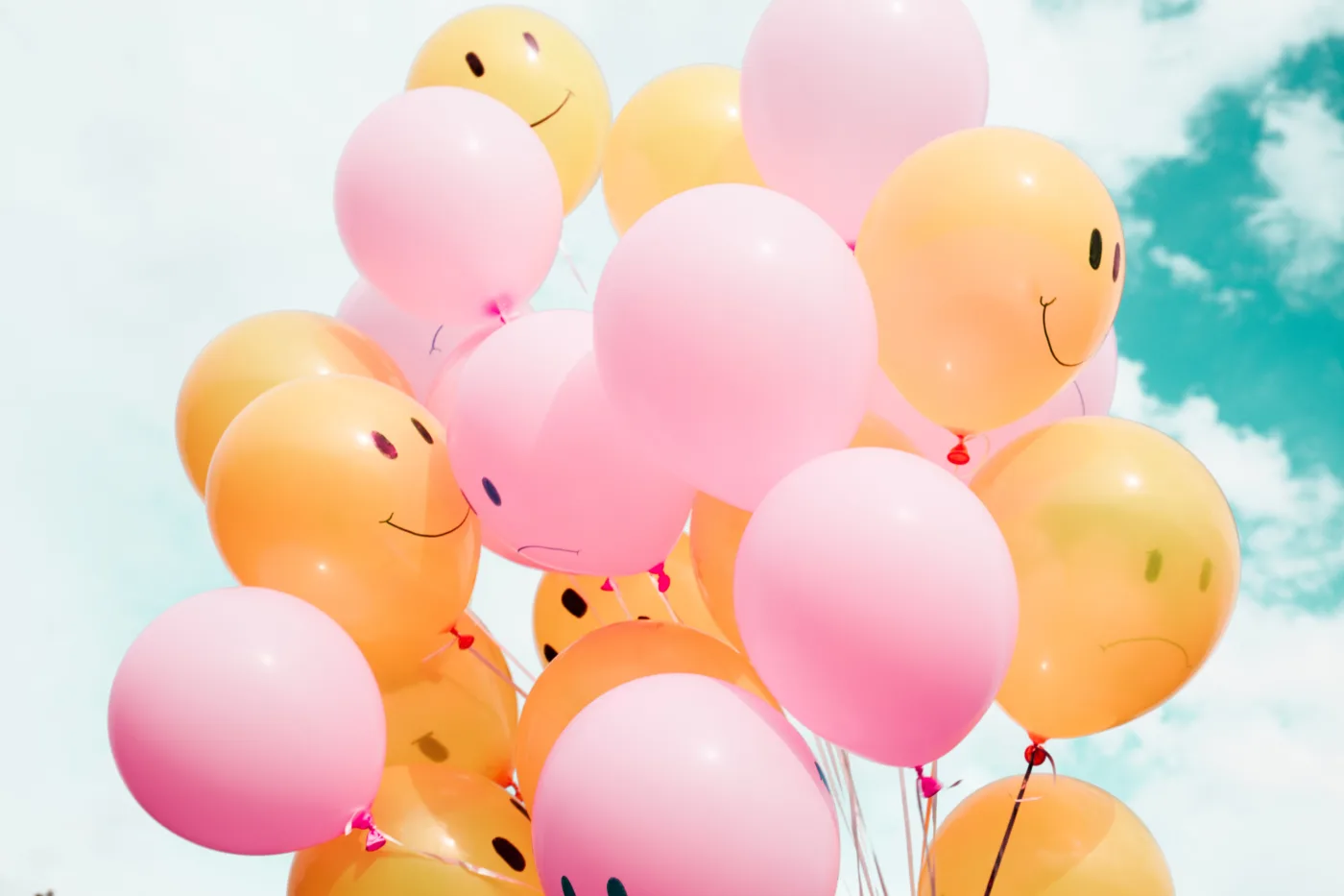 Lernmotivation – Ein Bündel von orangen und rosa Luftballons mit aufgemalten Gesichtern vor einem blauen Himmel mit Wolken.