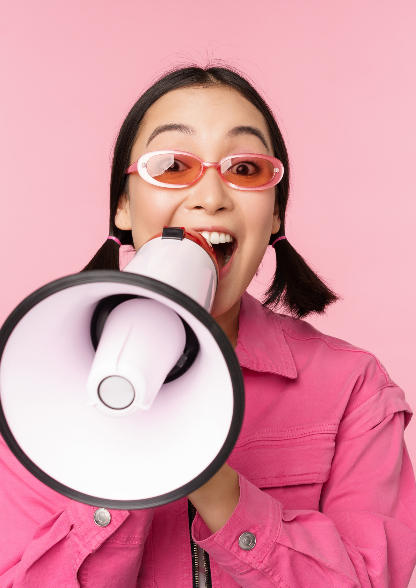 Mädchen in pinker Kleidung und Sonnenbrille spricht selbstbewusst durch ein Megafon vor rosa Hintergrund.