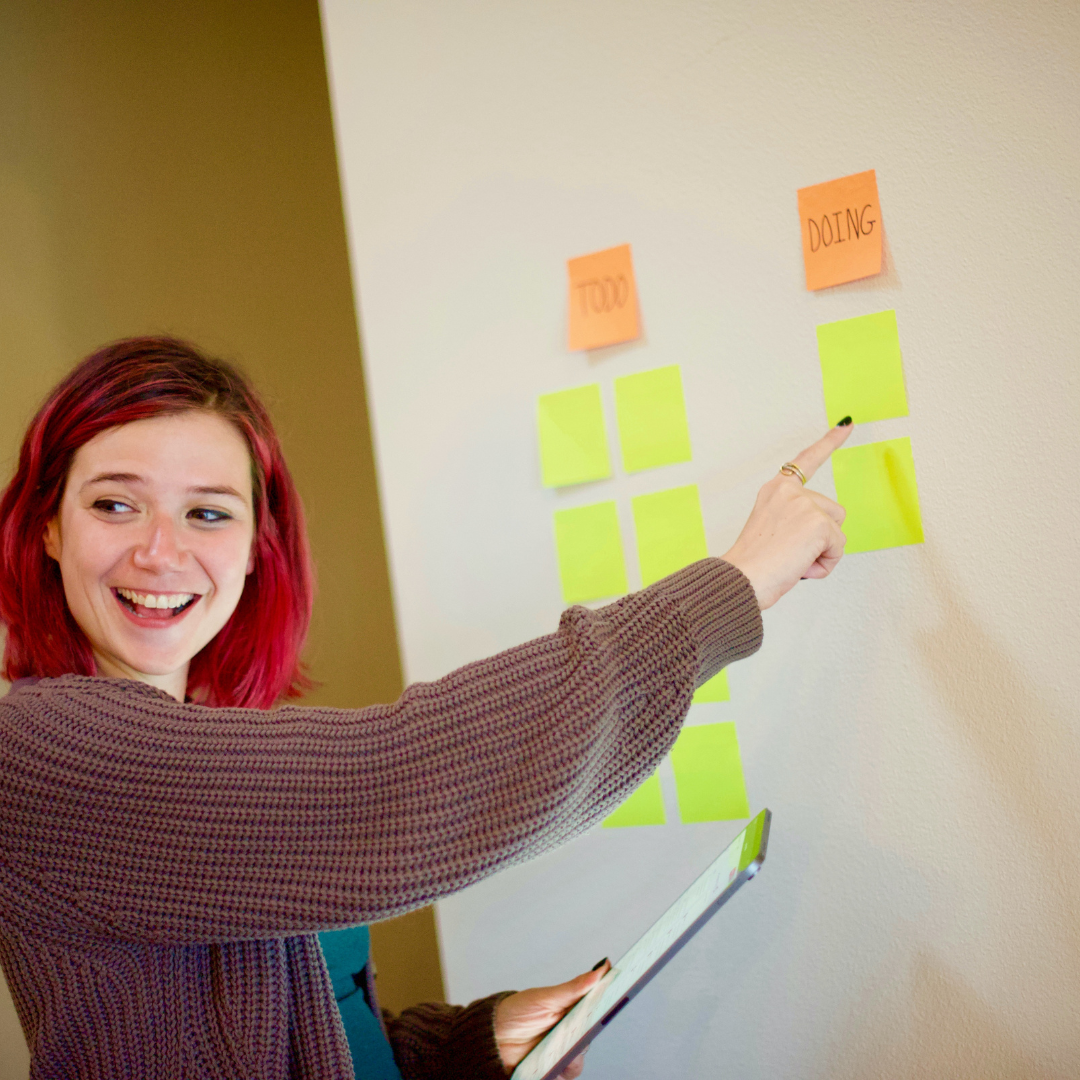 Eine lächelnde Frau mit roten Haaren platziert Post-its auf einem Whiteboard.