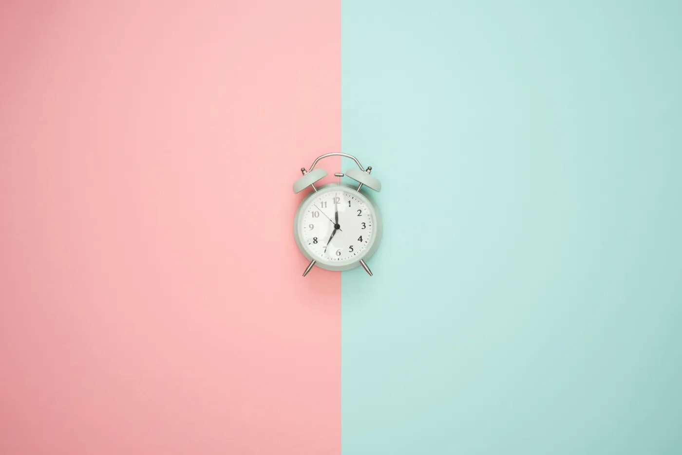 Ein klassischer Wecker steht in der Mitte einer zweigeteilten Fläche, die zur Hälfte rosa und zur anderen Hälfte hellblau gefärbt ist, und symbolisiert Zeitmanagement und Balance.