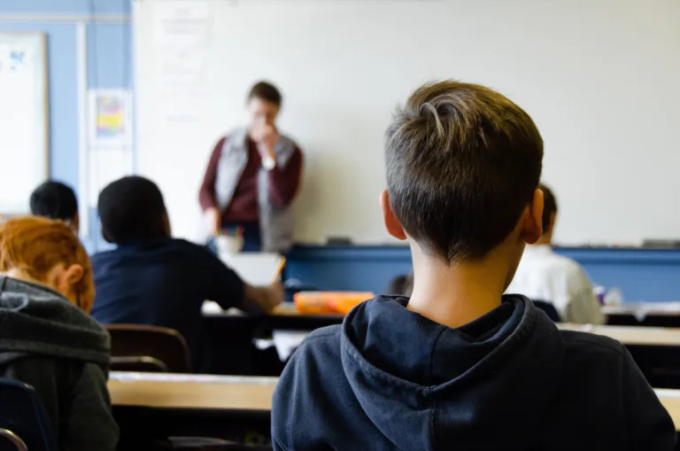 Ein Schüler mit Prüfungsangst in einem Klassenzimmer sitzt mit dem Rücken zur Kamera, während er auf einen Lehrer im Hintergrund blickt, der unscharf am Lehrerpult steht.