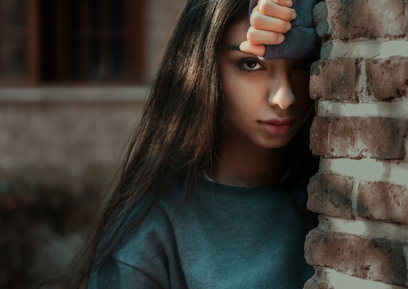 Eine junge Frau lehnt nachdenklich, wegen Prüfungsangst, an einer Backsteinmauer, ihr Gesicht teilweise verdeckt, während sie mit einer Hand über ihr Auge blickt, was ein Gefühl von Nachdenklichkeit oder Neugier ausdrückt.