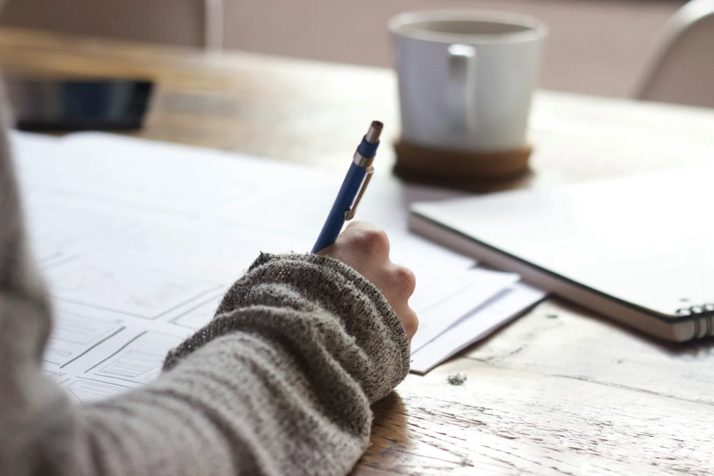 Eine Person bei der Prüfungsvorbereitung in einem gestrickten Pullover schreibt Notizen oder zeichnet in ein großes Papierblatt auf einem Holztisch, neben einer Tasse Kaffee und einem Clipboard.
