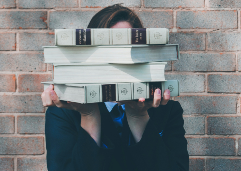 Eine Studentin verbirgt ihr Gesicht hinter einem Stapel dicker Enzyklopädien.