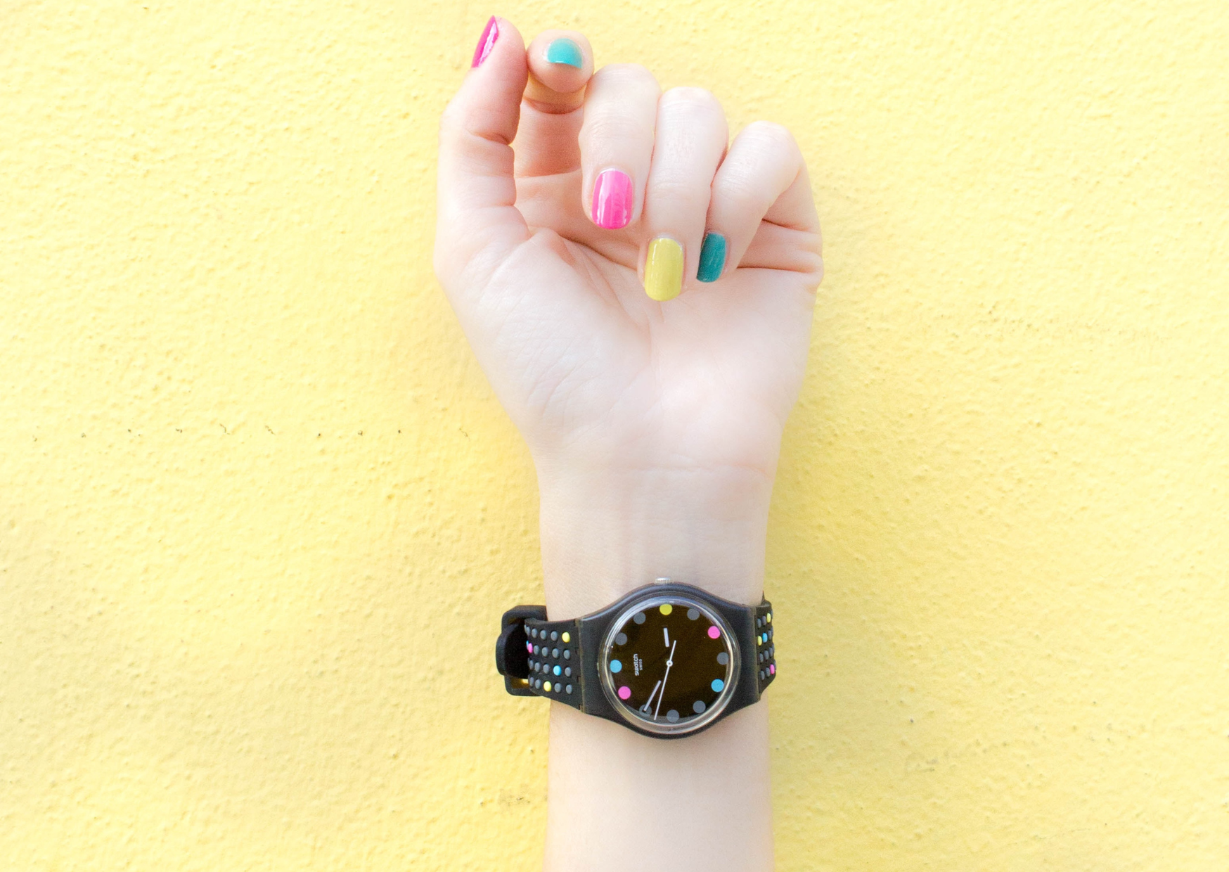 Eine erhobene Hand mit bunten Fingernägeln trägt eine stilvolle Armbanduhr vor einem gelben Hintergrund, der Fröhlichkeit und Zeitbewusstsein verkörpert.
